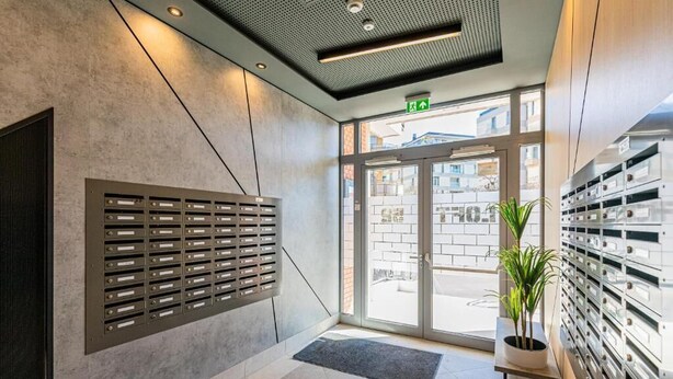 A modern lobby with a metal mail slot wall, a glass door entrance, and a potted plant.