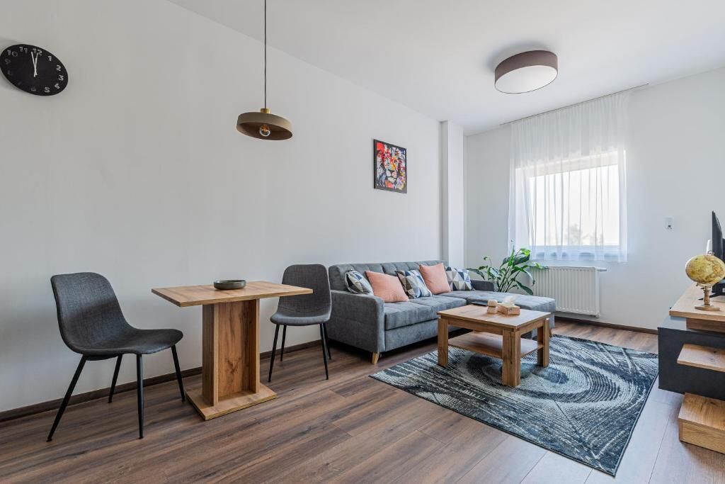 Ein modernes Wohnzimmer mit einer Couch, zwei Sesseln, einem Couchtisch, einem Esstisch, einer Uhr, einem Gemälde und einem Fenster mit Vorhängen.