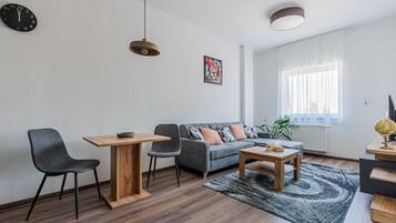 Ein modernes Wohnzimmer mit einer Couch, zwei Sesseln, einem Couchtisch, einem Esstisch, einer Uhr, einem Gemälde und einem Fenster mit Vorhängen.