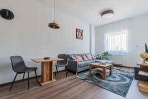 Un salon moderne comprenant un canapé, deux fauteuils, une table basse, une table à manger, une horloge, un tableau et une fenêtre avec des rideaux.