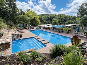 A swimming pool with a stone border, surrounded by lounge chairs and umbrellas, with a view of a lake and trees in the background.