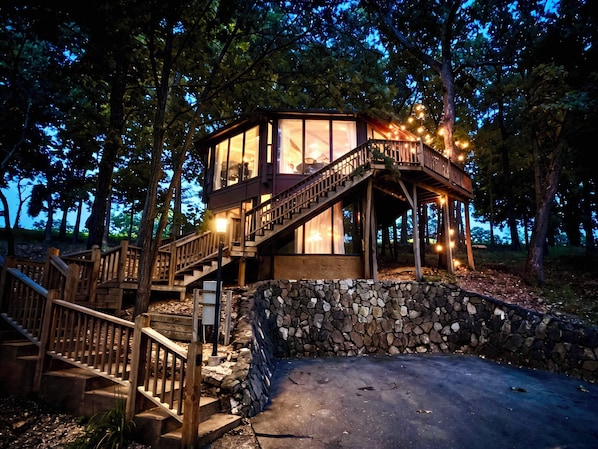A wooden cabin with a stone wall, wooden stairs, and a deck with string lights.