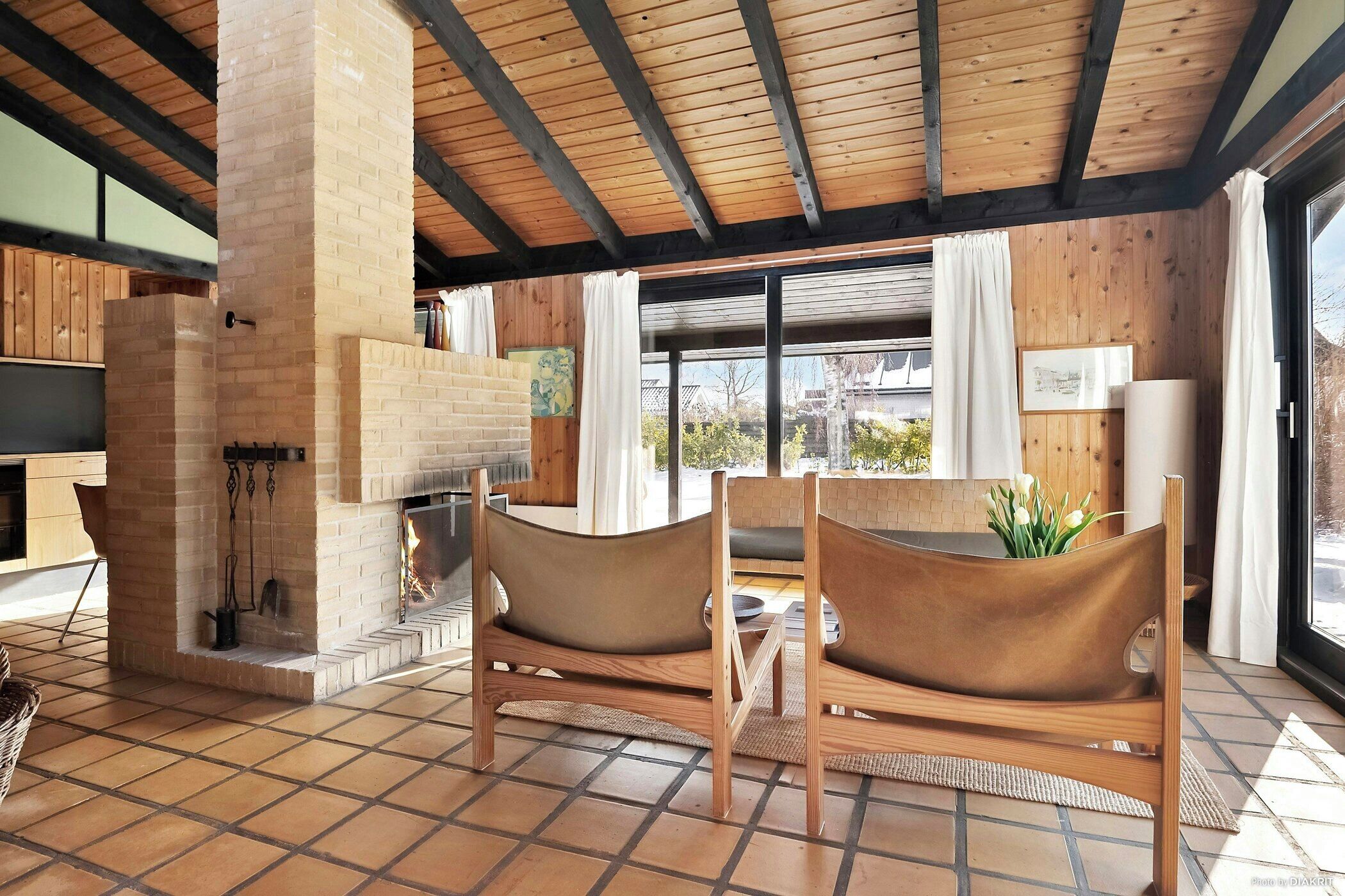 Een kamer met een open haard van baksteen, houten stoelen, een groot raam en uitzicht op buiten.