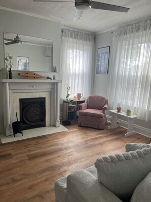 Een woonkamer met een open haard, een plafondventilator, een roze fauteuil en een witte bank.