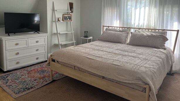 Une chambre à coucher avec un lit, un téléviseur posé sur une commode, une échelle et une fenêtre ornée de rideaux légers.
