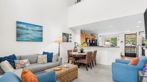 Un salon moderne avec un canapé gris, des fauteuils bleus, une table à manger avec des chaises, et une cuisine en arrière-plan.