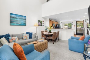 A modern living room with a grey sofa, blue armchairs, a dining table with chairs, and a kitchen in the background. - Alii Cove#EE22: Top Floor unit with Loft, Ac, Ocean Views, Sleeps 6! (Kailua Kona)