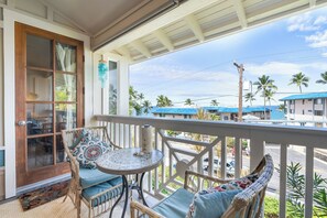 A balcony with wicker furniture, a small round table, and a view of a coastal area with buildings and palm trees. - Alii Cove#EE22: Top Floor unit with Loft, Ac, Ocean Views, Sleeps 6! (Kailua Kona)