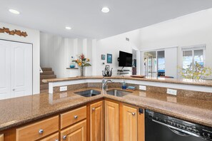 A kitchen with granite countertops, wooden cabinets, a dishwasher, and a TV mounted on the wall. - Alii Cove#EE22: Top Floor unit with Loft, Ac, Ocean Views, Sleeps 6! (Kailua Kona)