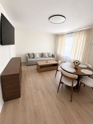 A modern living room with a sofa, a coffee table, a TV mounted on the wall, and a dining table with chairs.