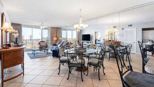 A spacious living area with a dining table, chairs, a TV, and a balcony with a view. - Terrace At Pelican Beach 602 2 Bedroom Home (Destin)