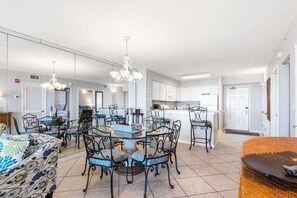 A modern kitchen with white cabinets, a dining area with a glass table and metal chairs, and a living area with a sofa and patterned throw. - Terrace At Pelican Beach 602 2 Bedroom Home (Destin)