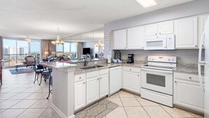 A modern kitchen with white cabinets, a central island, and a dining area with a view. - Terrace At Pelican Beach 602 2 Bedroom Home (Destin)