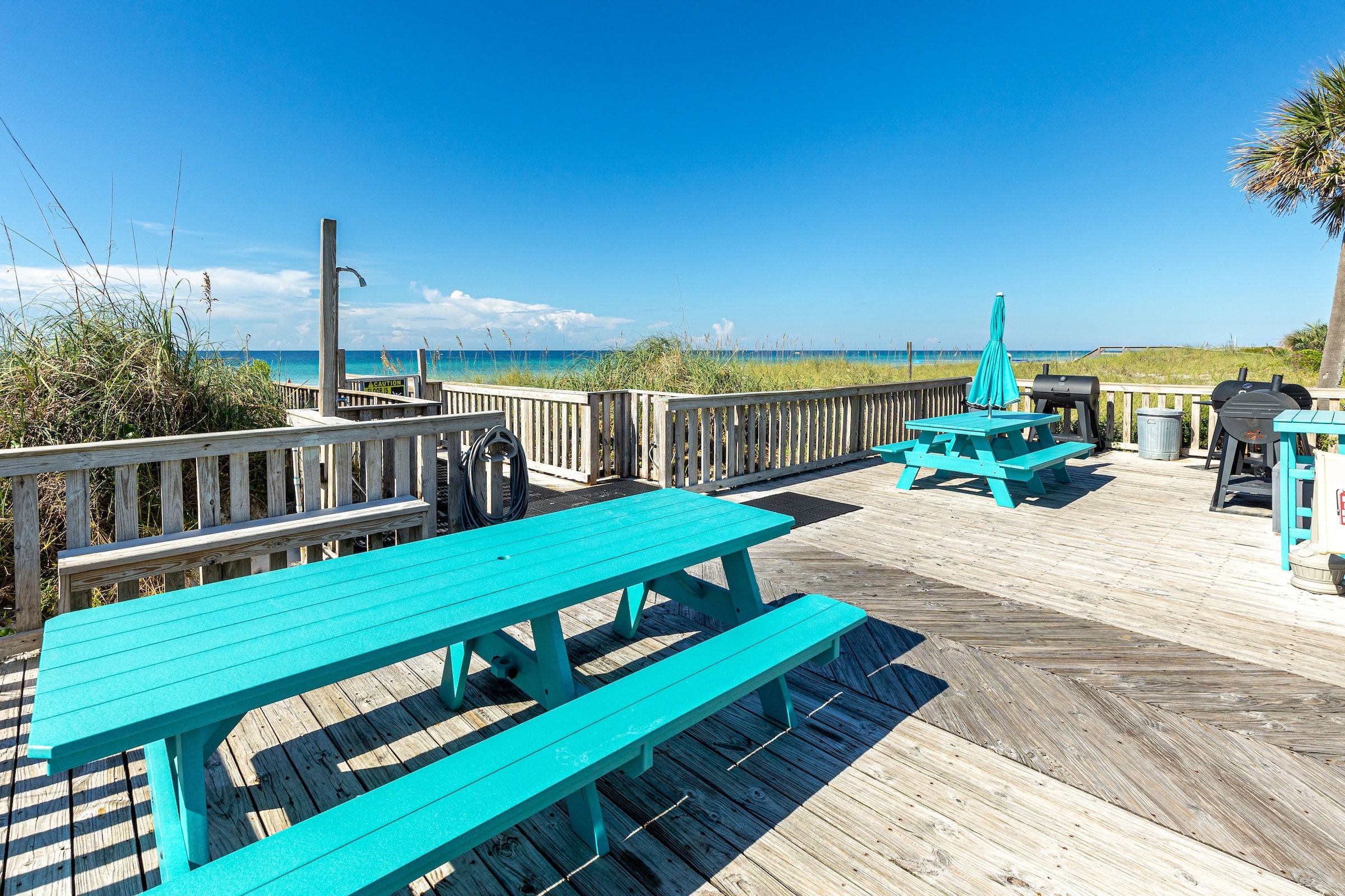 A wooden deck with turquoise picnic tables and a barbecue grill, overlooking a beach.