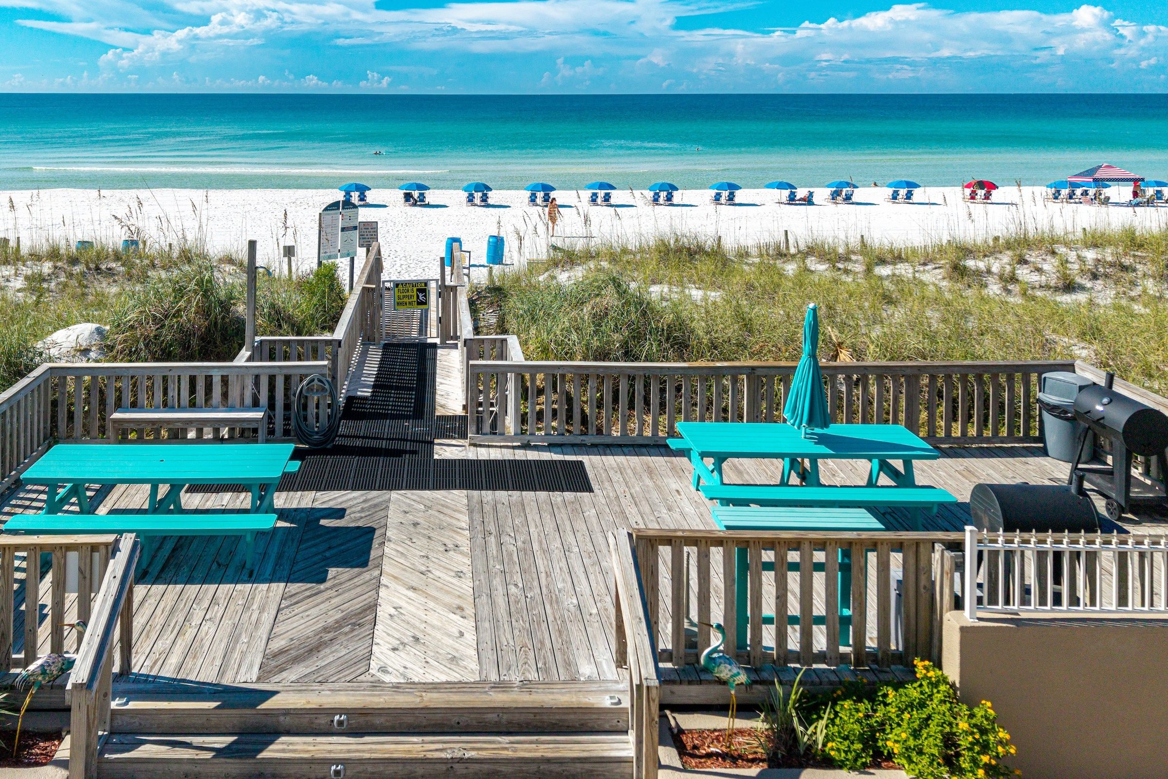 A beachside deck with picnic tables and a barbecue grill, overlooking a sandy beach with umbrellas.