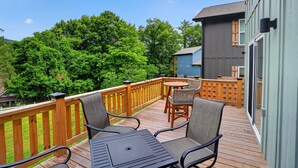 A wooden deck with outdoor furniture, a view of a forest, and a building with a dark exterior. - Mountain Views Basecamp West Jefferson 2 Bedroom Home (West Jefferson)