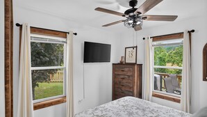 A bedroom with a bed, a ceiling fan, a TV, and windows with curtains. - Mountain Views Basecamp West Jefferson 2 Bedroom Home (West Jefferson)