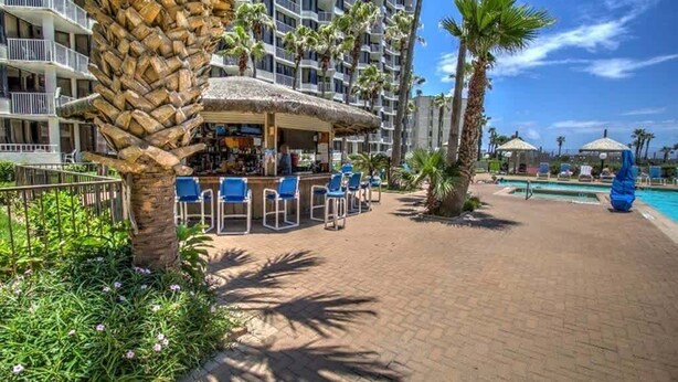 Un hotel con área de bar, mesas al aire libre, piscina y palmeras.