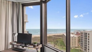Una habitación con un ventanal grande, un escritorio con computadora y vista a la playa y edificios.