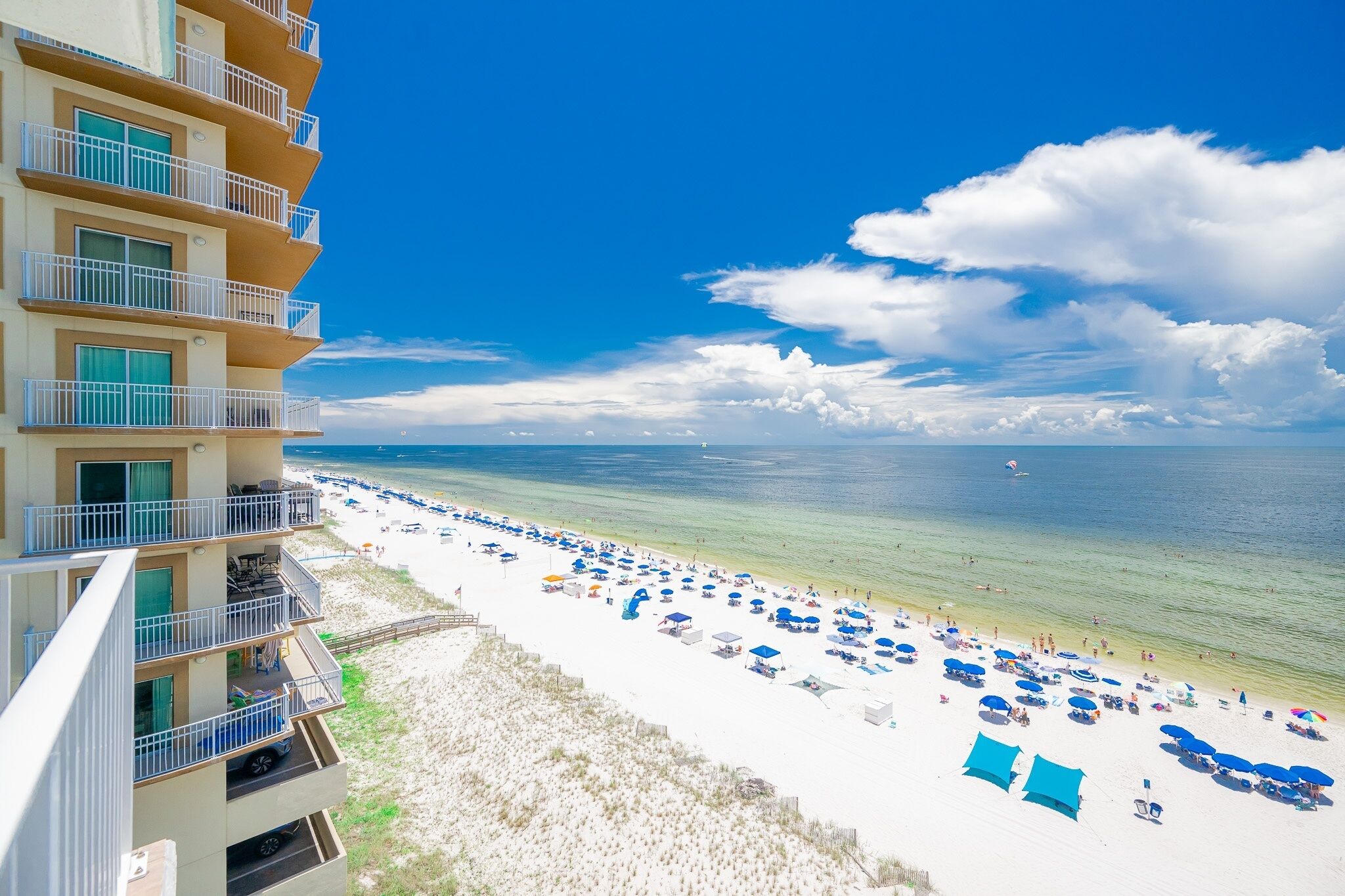 A beachfront hotel with multiple balconies, a sandy beach with umbrellas, and a clear view of the ocean.