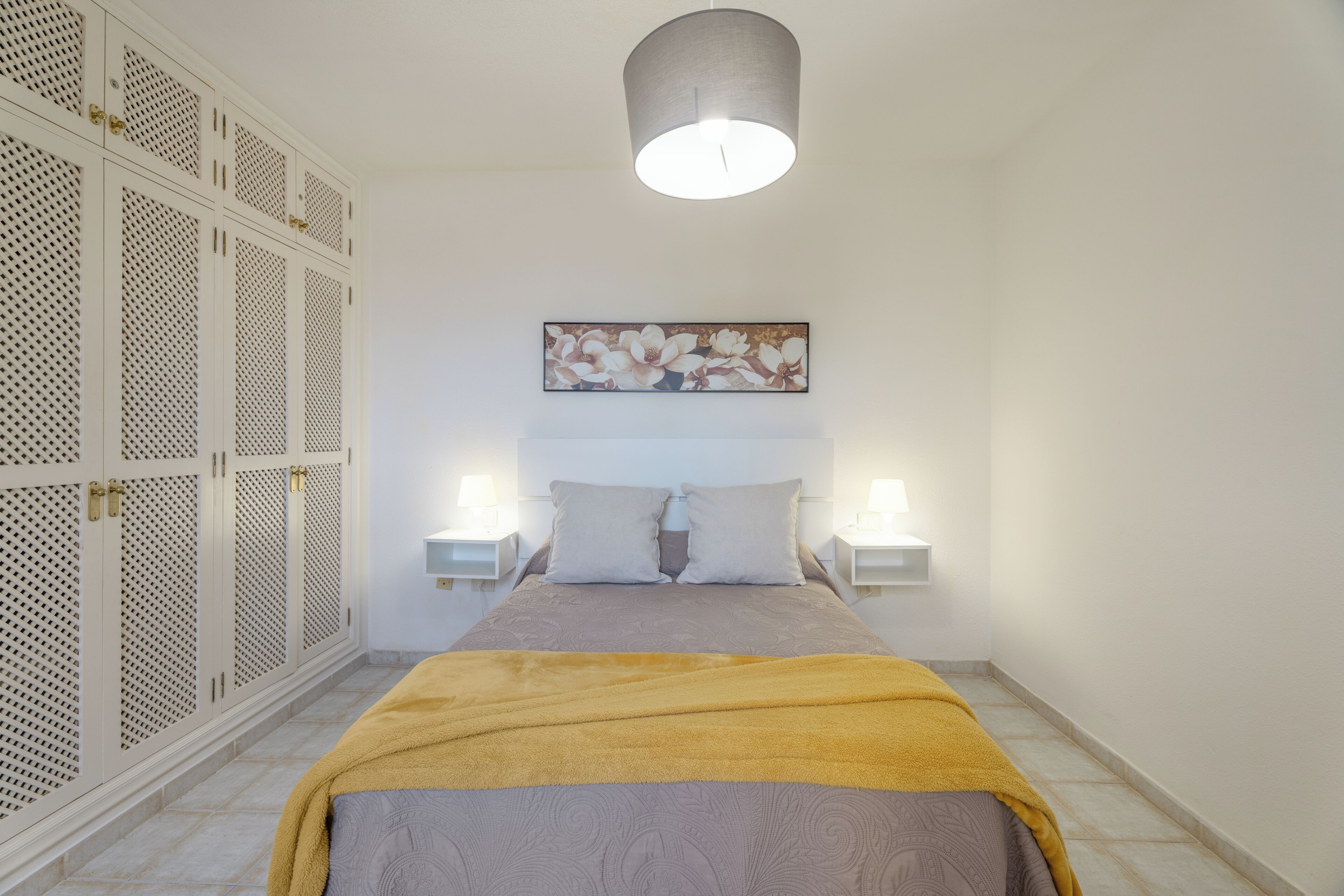 Un dormitorio con cama, cabecera, dos mesitas de noche, lámpara de pared, luz de techo y armario con diseño perforado.