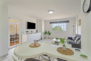 Uma sala de estar moderna com televisão de tela plana, um sofá, uma mesa de jantar com cadeiras e uma área de cozinha com geladeira e plantas em vasos.