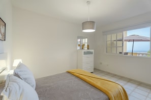 Quarto com cama, mesa de cabeceira, abajur, cômoda, janela com um guarda-chuva e vista para o mar.