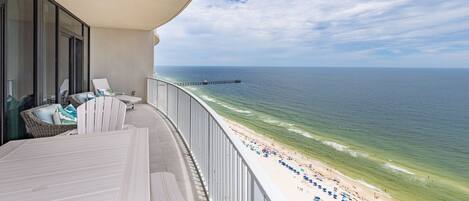A balcony with a view of the beach and ocean, a pier in the distance, and outdoor seating.