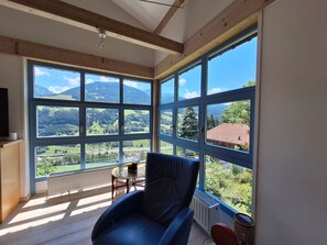 Ein Zimmer mit großen Fenstern, einem Sessel, einer Heizung und einem Fernseher.