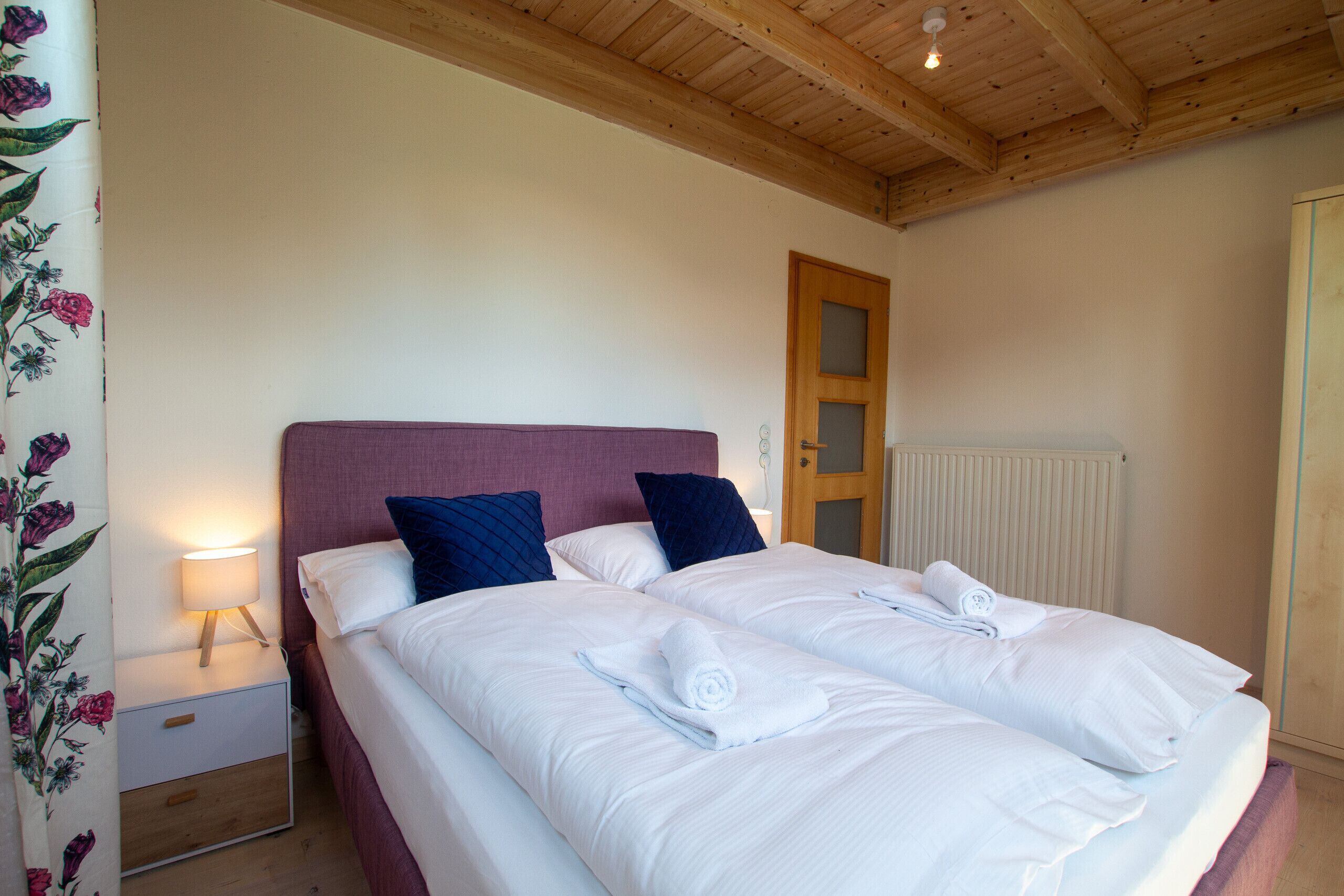 Una cama doble con sábanas blancas, dos almohadas, una mesita de noche con lámpara y techo de madera.