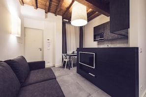 Ein modernes Wohnzimmer mit einem dunkelgrauen Sofa, einem Essbereich mit Tisch und Stühlen, einer Küche mit schwarzen Schränken und einem Fernseher, sowie einer Holzbalkendecke.