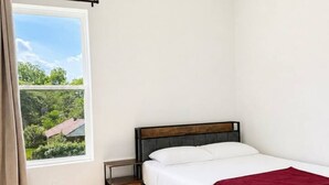 Ein Einzelbett mit Holzrahmen, ein Nachttisch, ein Fenster mit Vorhängen und Blick ins Grüne.
