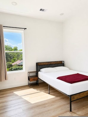A single bed with a wooden frame, a bedside table, a window with curtains, and a view of greenery outside. - Modern 3BR Near UH Downtown Stadiums (Houston)