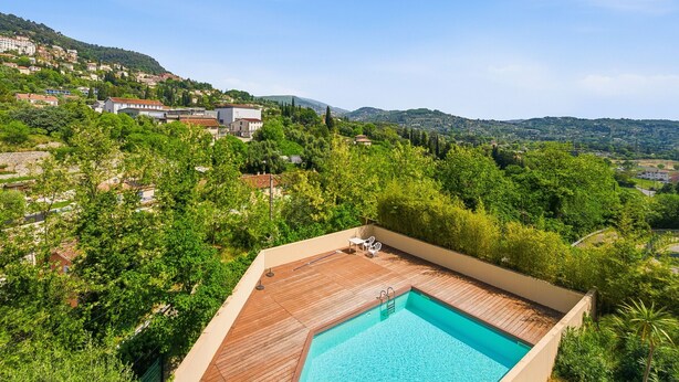 Una piscina sul tetto con un solarium in legno, circondata da vegetazione lussureggiante e vista su una cittadina collinare.