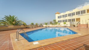 Una piscina con plataforma de baldosas, escalera metálica y un edificio con varias ventanas al fondo.