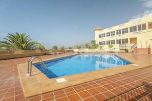 A swimming pool with a tiled deck, a metal ladder, and a building with multiple windows in the background. - Apartment Balcon del Atlantico, Costa Adeje (Playa de las Américas)