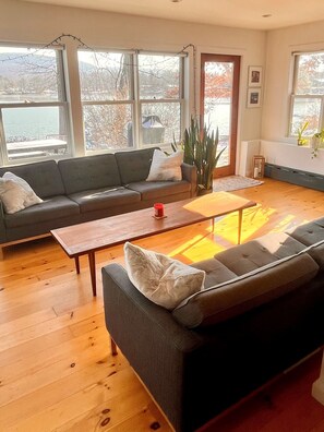 Sala de estar con sofá, mesa de centro y ventanas amplias con vista a árboles y una masa de agua.