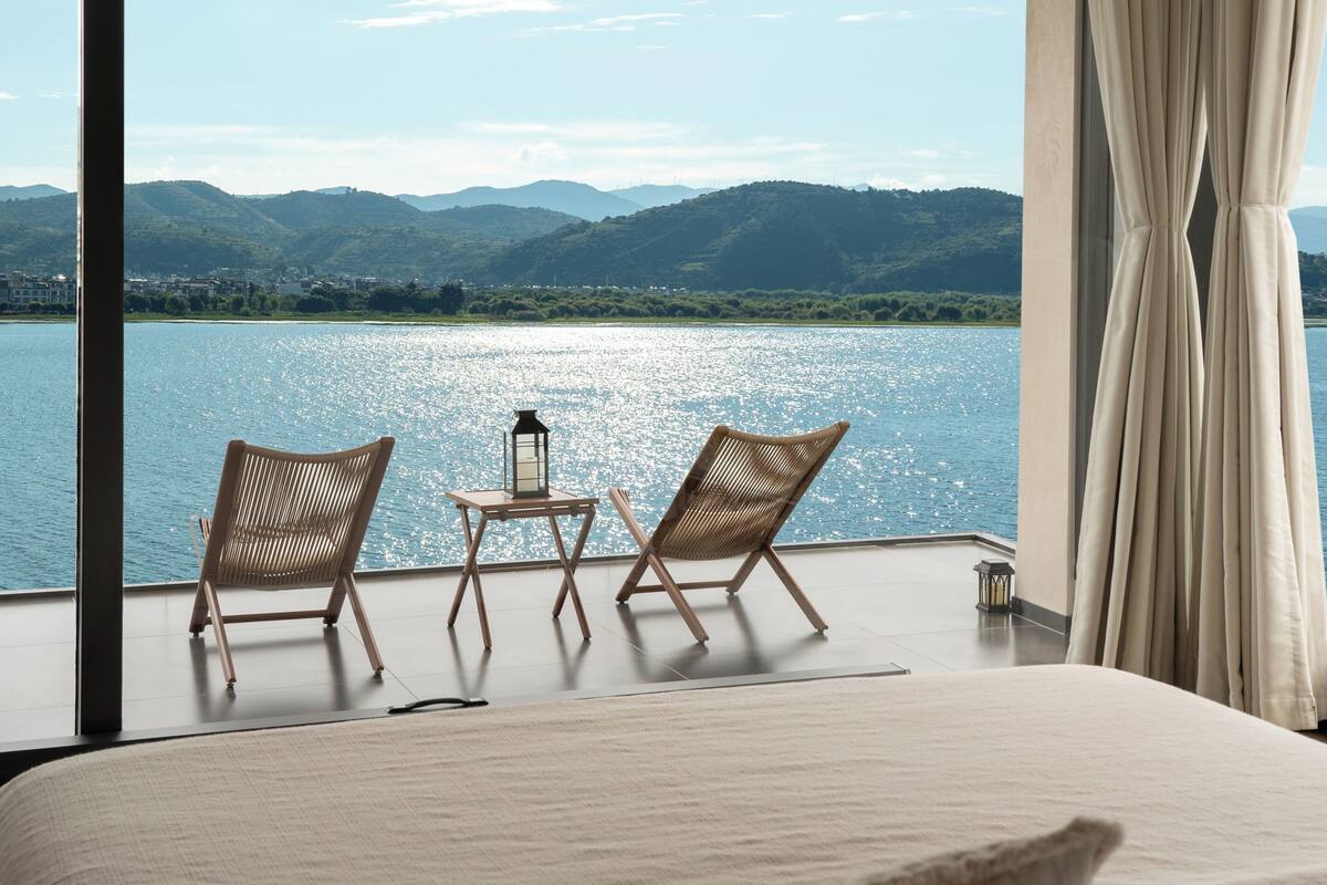 Un dormitorio con una cama grande, dos sillas de mimbre, una mesita y vistas a un lago y montañas.