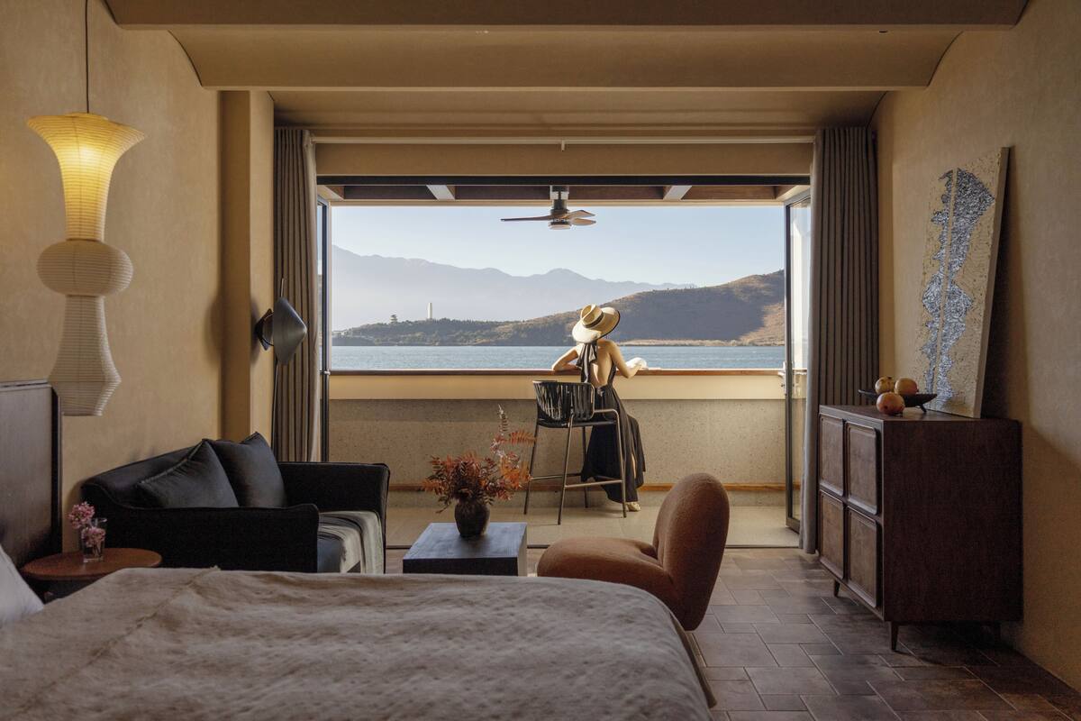 Habitación de hotel con cama, sofá, escritorio con un florero, silla, armario y vistas al mar y a las montañas.