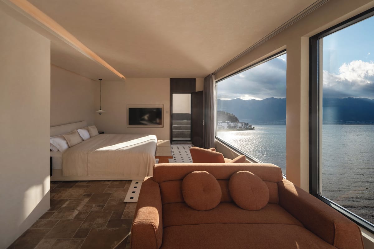 Una habitación moderna con cama, sofá y ventanas amplias con vista a un cuerpo de agua y montañas.