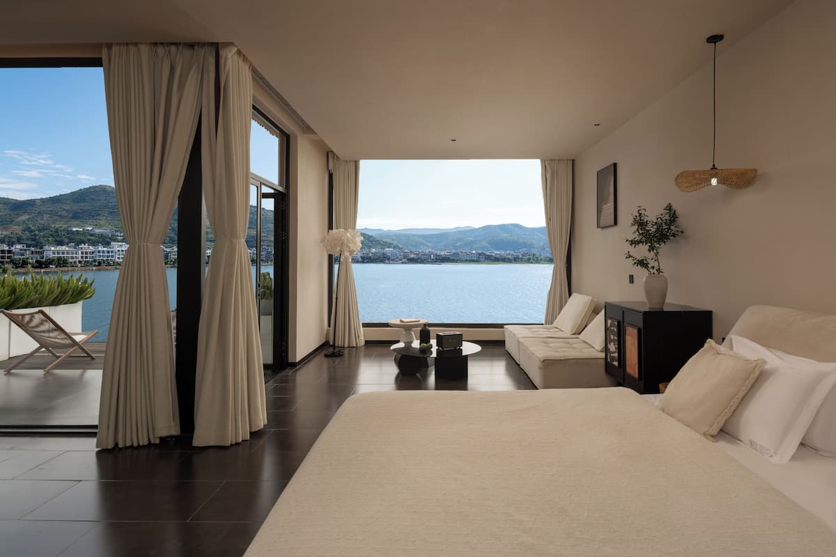 Una habitación de hotel moderna con una cama grande, un sofá, una mesita y vistas a un lago y montañas.