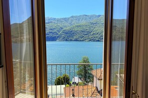Blick durch ein Fenster auf einen See, umgeben von Hügeln, mit einem Balkon und Gebäuden mit roten Ziegeldächern.