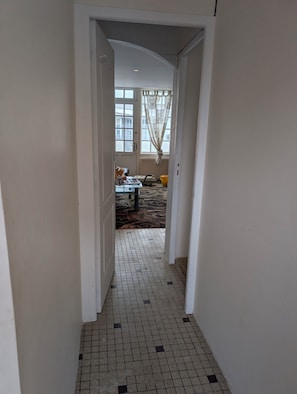 Un couloir carrelé mène à une pièce où se trouvent un lit, une table et une fenêtre avec des rideaux.