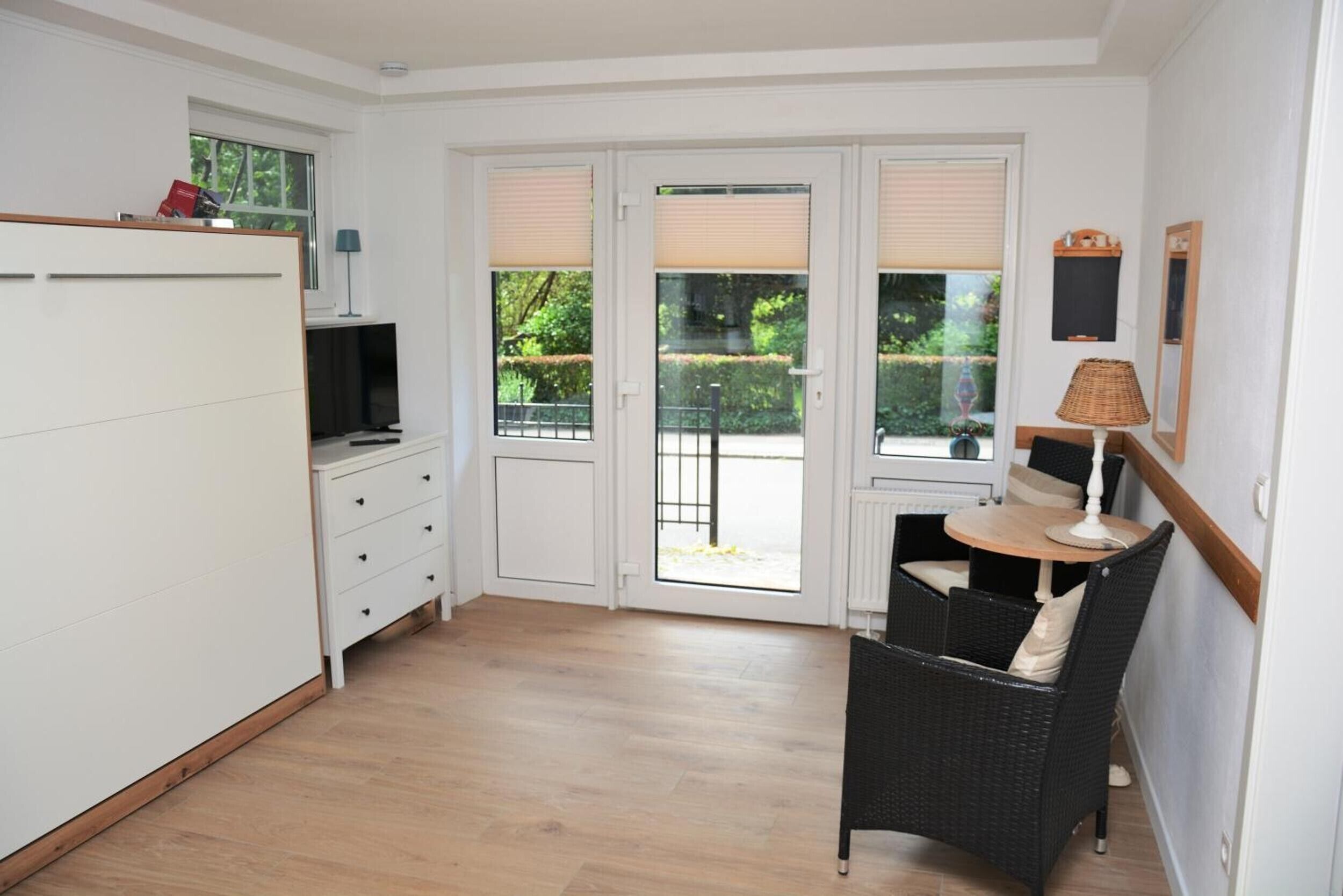 Ein Zimmer mit einem weißen Kleiderschrank, einem Fernseher, einem Korbstuhl, einem Tisch und Blick durch eine Glastür.