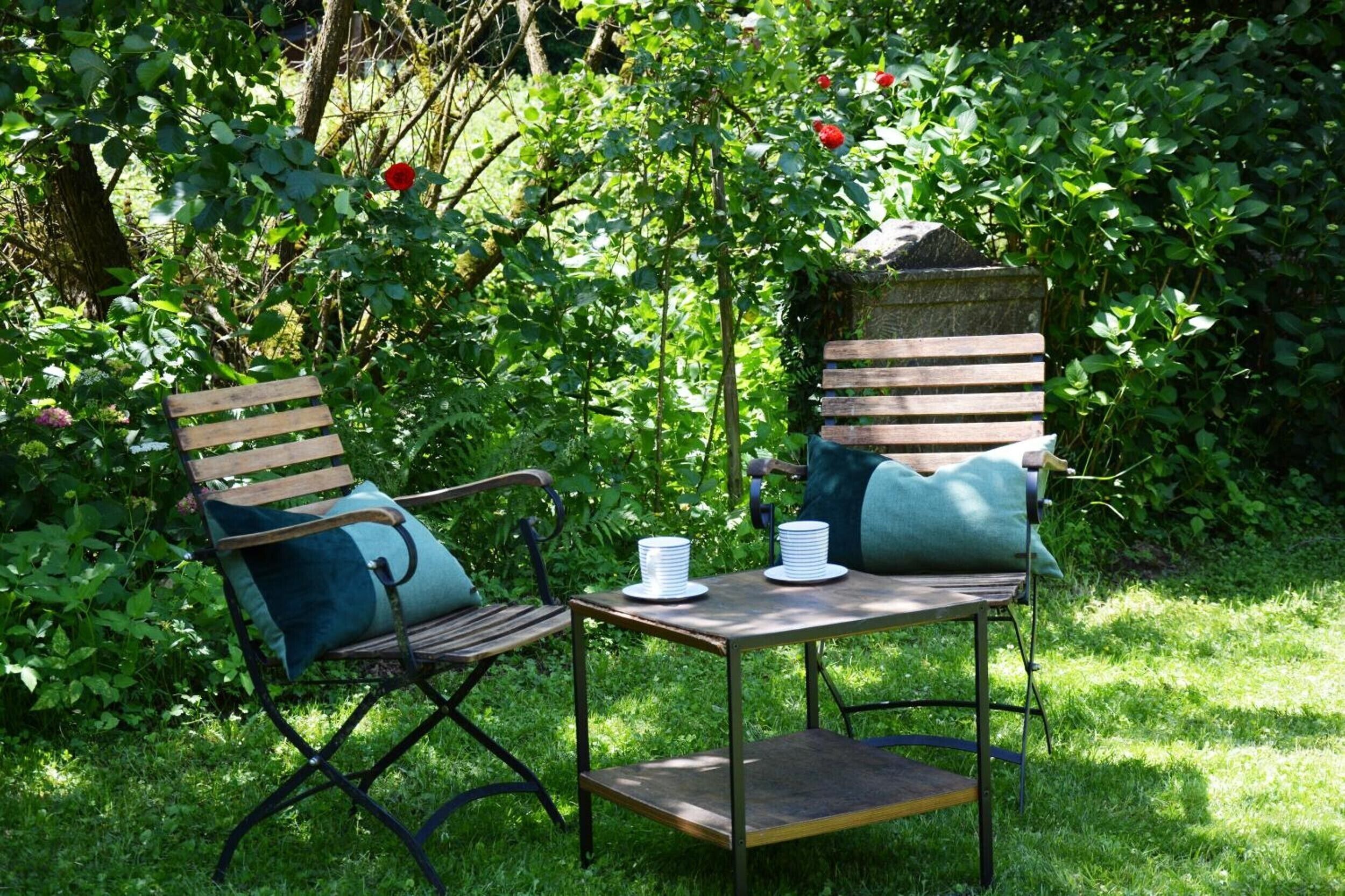 A garden with two wooden chairs and a small table, surrounded by lush greenery and red flowers.