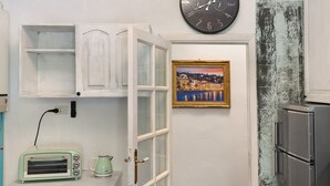 Une cuisine avec des meubles blancs, un grille-pain vintage, une horloge et un tableau représentant un paysage urbain encadré.