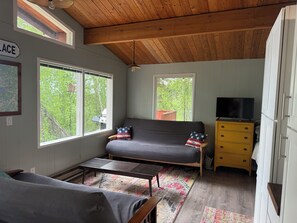 Ein gemütliches Zimmer mit einer Couch, einem Fernseher, einer Kommode und einem Fenster mit Blick auf Bäume.