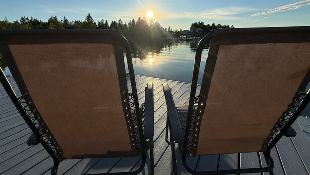 Een houten terras met twee stoelen gericht op een rustig meer en een ondergaande zon.
