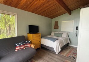 Ein Schlafzimmer mit einem Bett, einem Schlafsofa, einer Holzkommode, einem Fernseher und einem Fenster mit Jalousien.