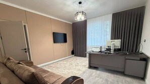 Una sala de estar moderna con un sofá, un televisor de pantalla plana, un escritorio con una computadora y un amplio ventanal con cortinas.